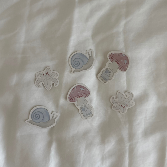 Six stickers displayed up close on a white background, featuring two snails, two mushroom characters, and two lilies (each design repeated twice).