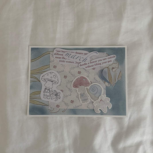 Inside an open envelope: 5x7 white lily print, 4x4 matching patterned print in soft blues, three matte stickers, journal and activity prompts, a mini mushroom coloring card, mini journaling cards, and three small lined paper hearts.