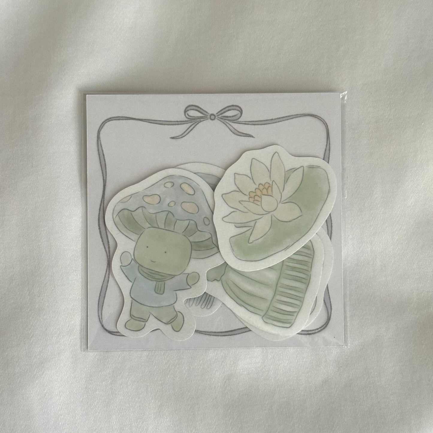 Five-piece sticker set with illustrated designs of a water lily, a mushroom character, a beanie, a scarf, and a mug, plus a 3x3 memo paper sheet, packaged in a clear sleeve on a white background.