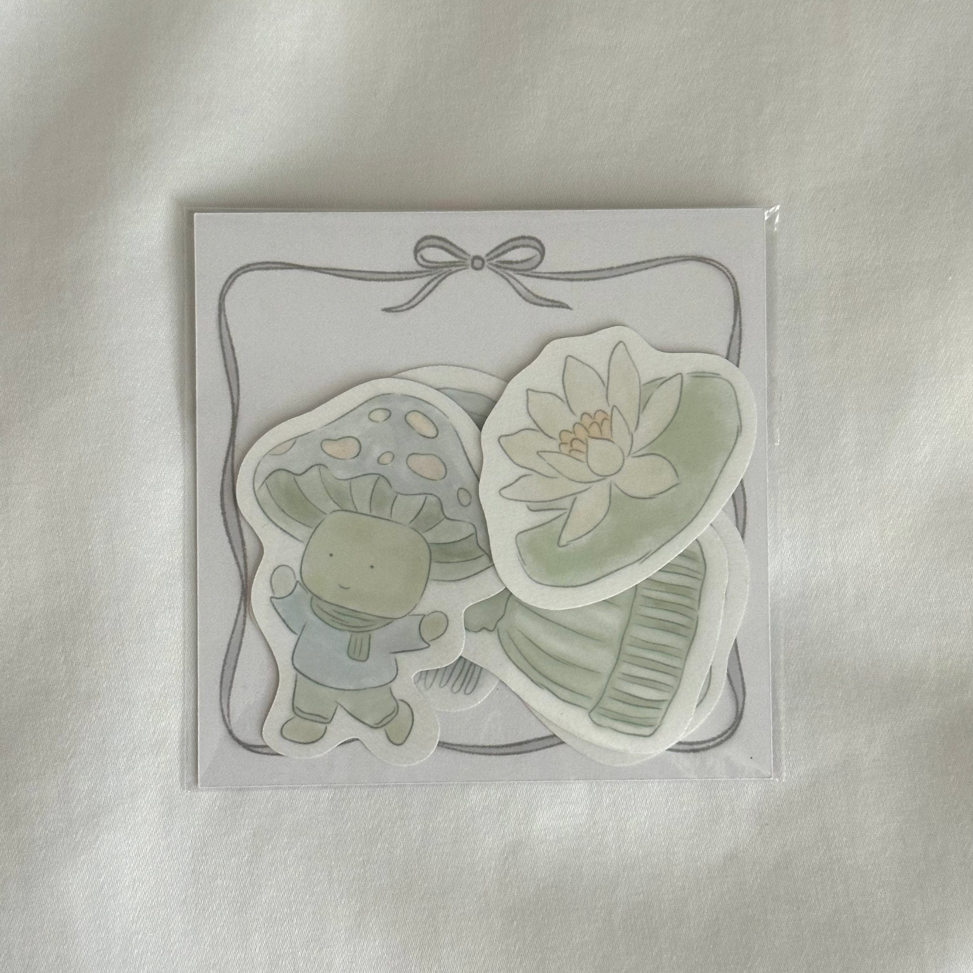 Five-piece sticker set with illustrated designs of a water lily, a mushroom character, a beanie, a scarf, and a mug, plus a 3x3 memo paper sheet, packaged in a clear sleeve on a white background.