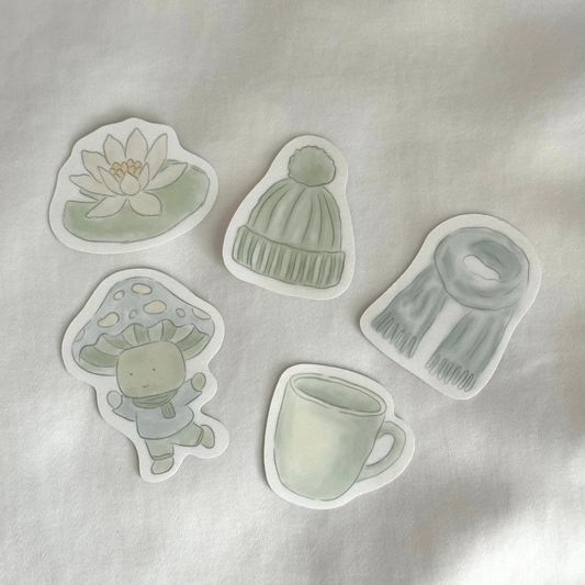 Six stickers displayed up close on a white background, featuring a water lily, a beanie, a scarf, a mushroom character, and a mug (all in soft sage green tones)