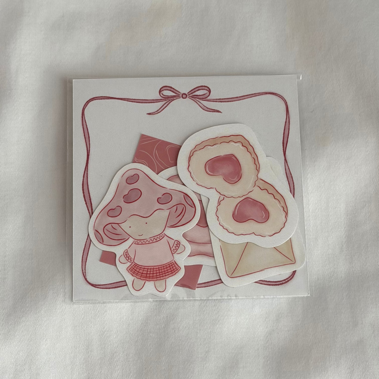 Five-piece sticker set with illustrated designs of a pair of cookies, mushroom character, mini art print, an envelope and a stamp seal, plus a 3x3 memo paper sheet, packaged in a clear sleeve on a white background.